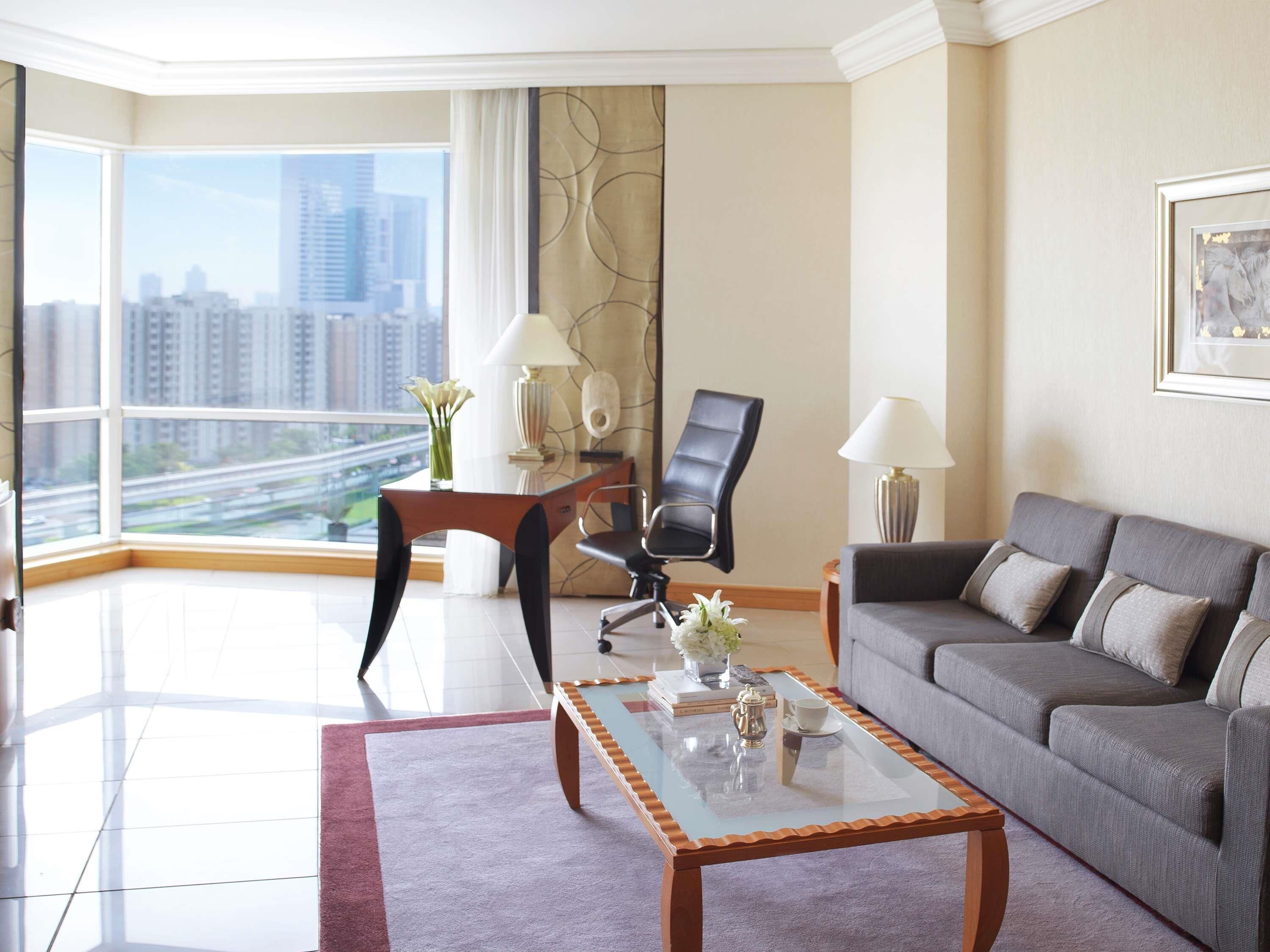 Una sala moderna con una mesa de centro de vidrio, un sofá gris, una silla de oficina negra y vistas a un paisaje urbano a través de ventanas grandes.