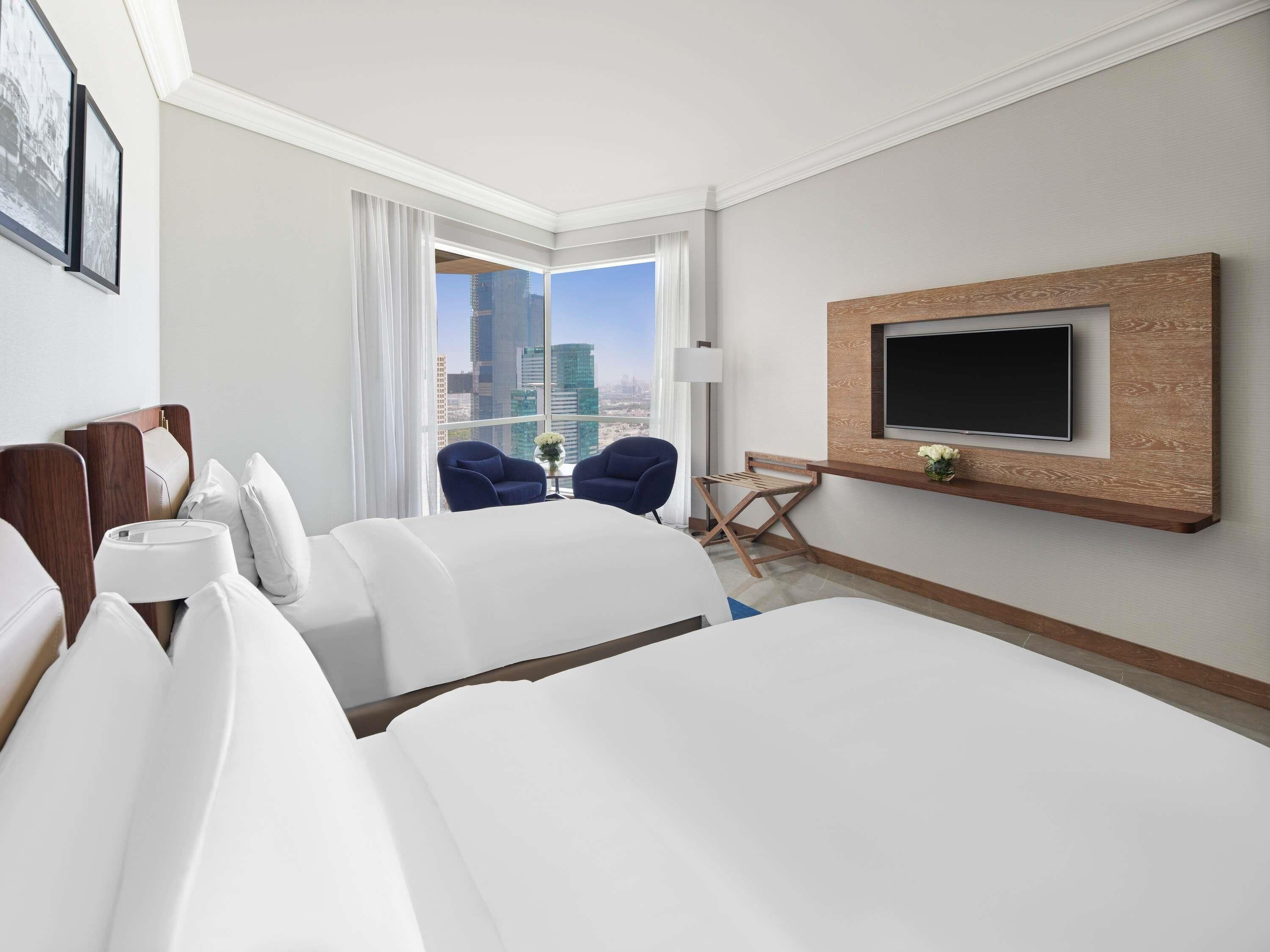 Ein Hotelzimmer mit einem großen Bett, zwei Sesseln, einem an einer Holzplatte montierten Fernseher und Blick auf die Stadt.