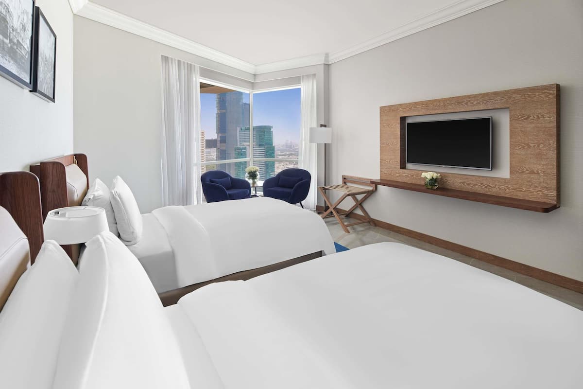 Ein Hotelzimmer mit einem großen Bett, zwei Sesseln, einem an einer Holzplatte montierten Fernseher und Blick auf die Stadt.