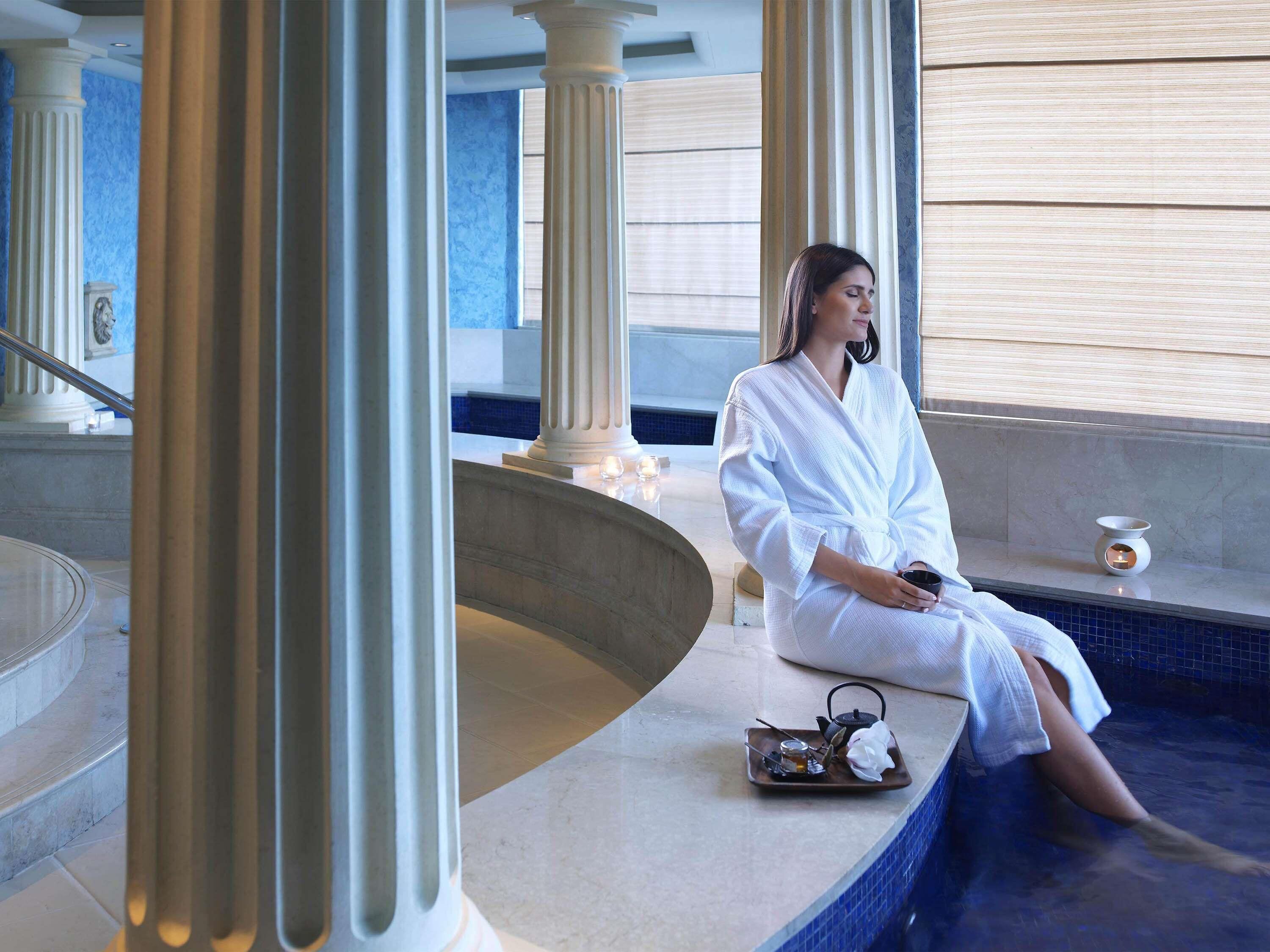 Una persona sentada junto a una piscina en un entorno de spa, con columnas y una pared de azulejos.