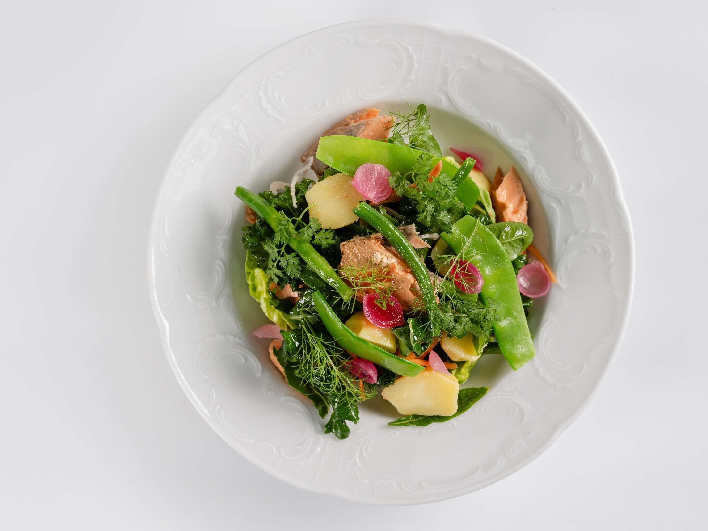 A white plate with a salad containing greens, potatoes, and salmon.