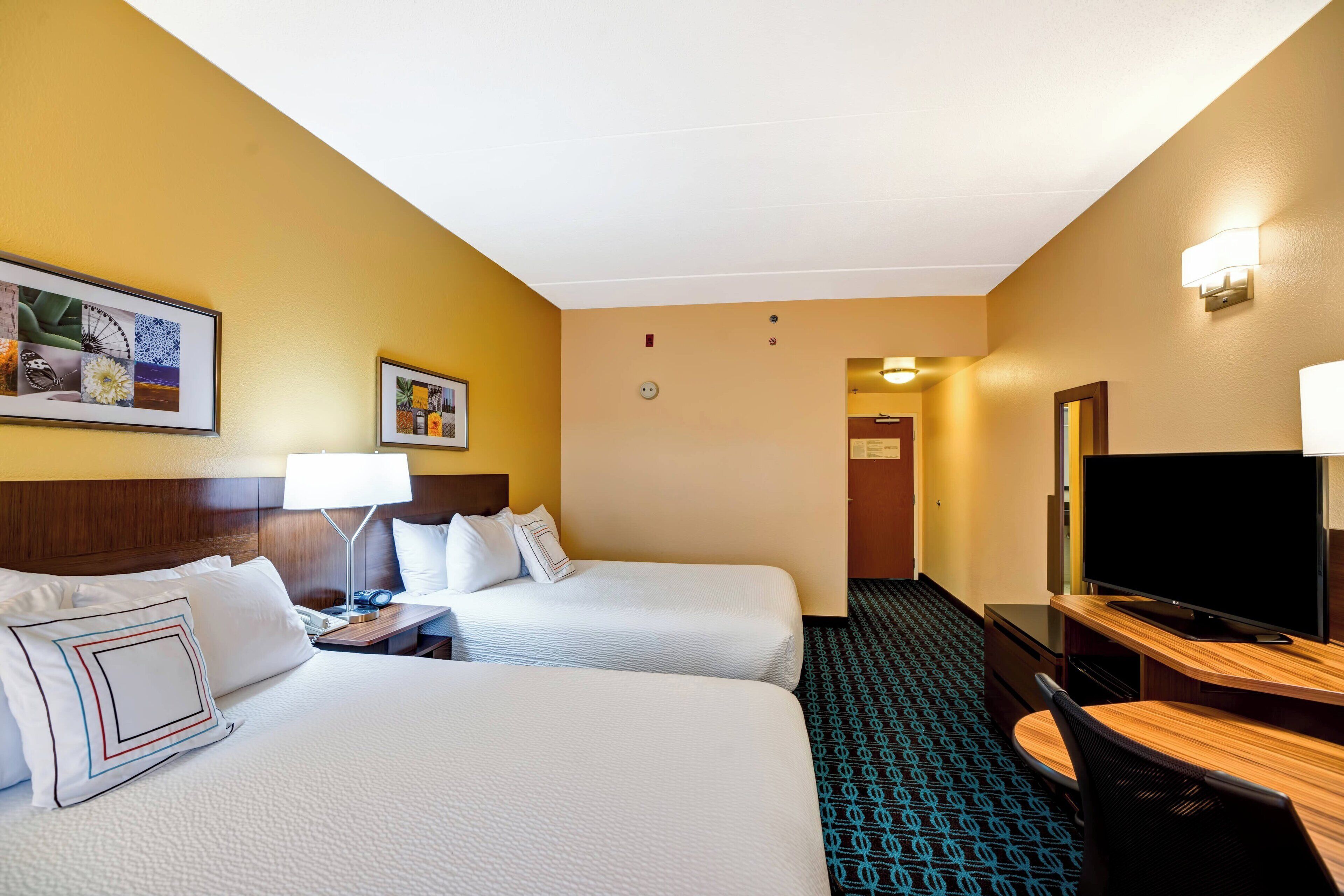 Quarto de hotel com duas camas, uma mesa de trabalho, uma televisão, um abajur e quadros nas paredes.