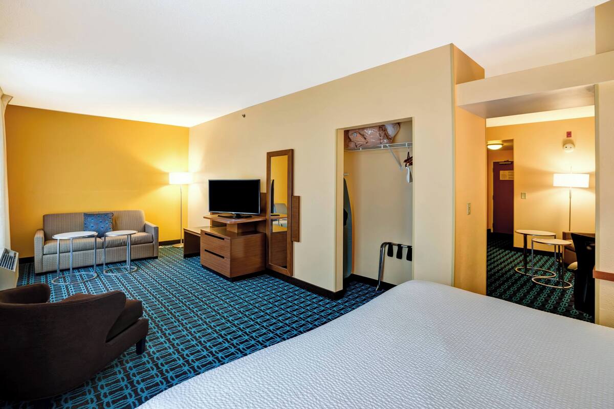 A hotel room with a bed, a sofa, a TV, a desk, and a bathroom visible through an open door.