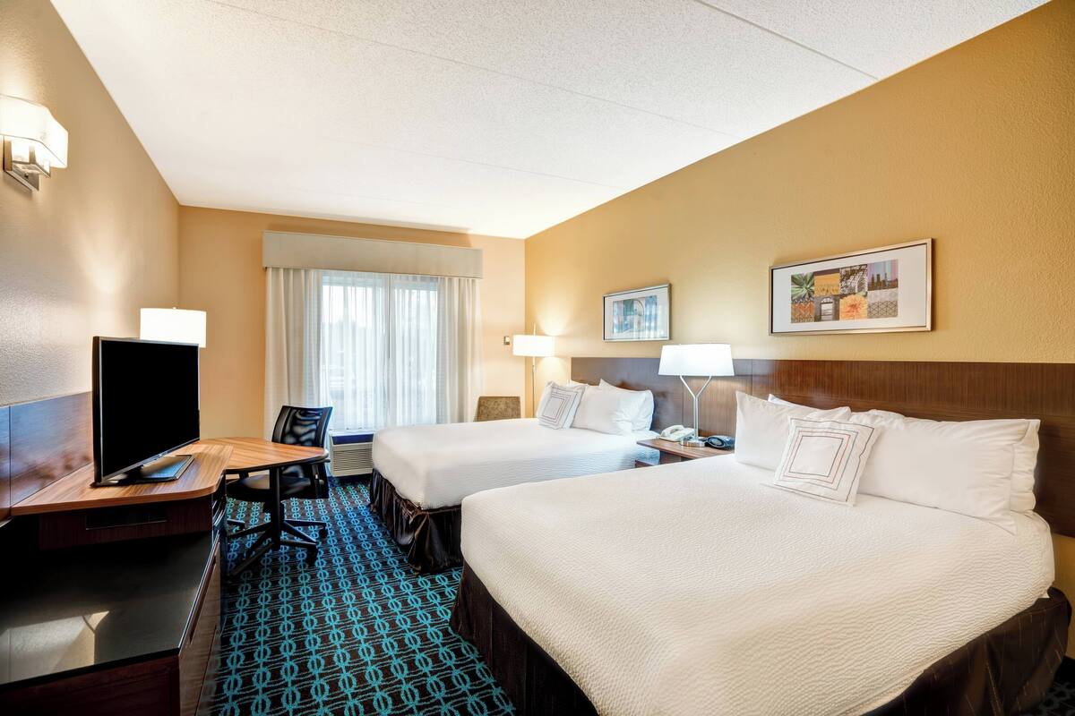 A hotel room with two beds, a desk with a flat-screen TV, a chair, and a patterned carpet.