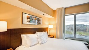 Ein Hotelzimmer mit einem großen Bett, einem gerahmten Bild an der Wand, einem Nachttisch mit einer Lampe und einem Fenster mit Blick ins Freie.