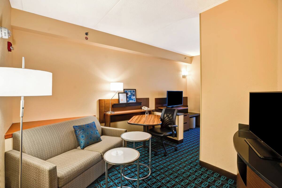A hotel room with a sofa, two small tables, a desk with a computer, a TV, and a lamp.