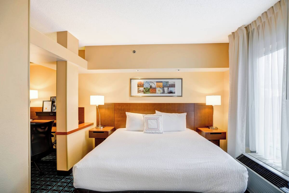 A hotel room with a large bed, two bedside tables, a desk with a chair, a headboard with a picture, and a window with curtains.
