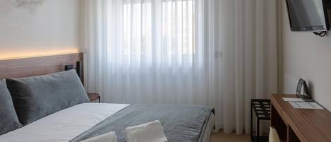 A hotel room with a bed, a desk, a TV, and a window with sheer curtains.