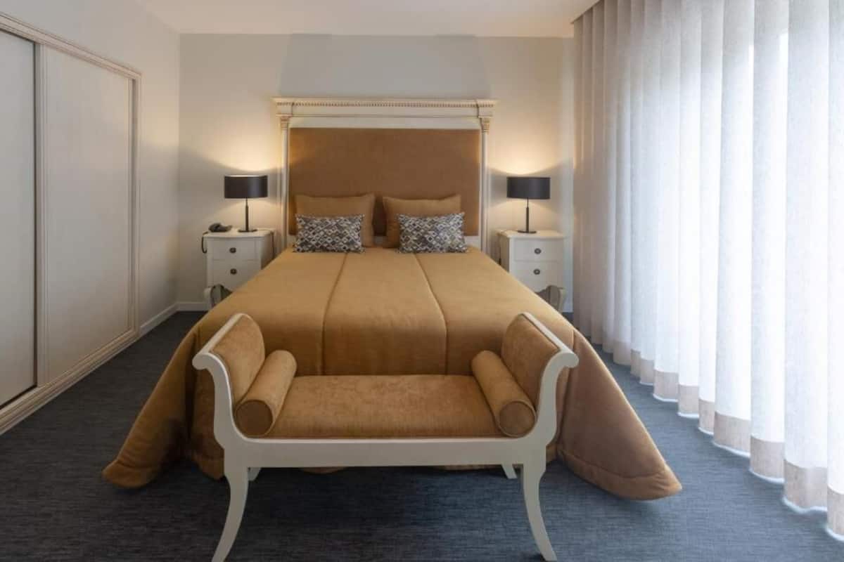 Habitación de hotel con una cama grande, dos mesitas de noche con lámparas, una silla blanca y un ventanal con cortinas.