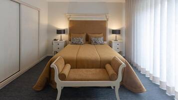 Habitación de hotel con una cama grande, dos mesitas de noche con lámparas, una silla blanca y un ventanal con cortinas.