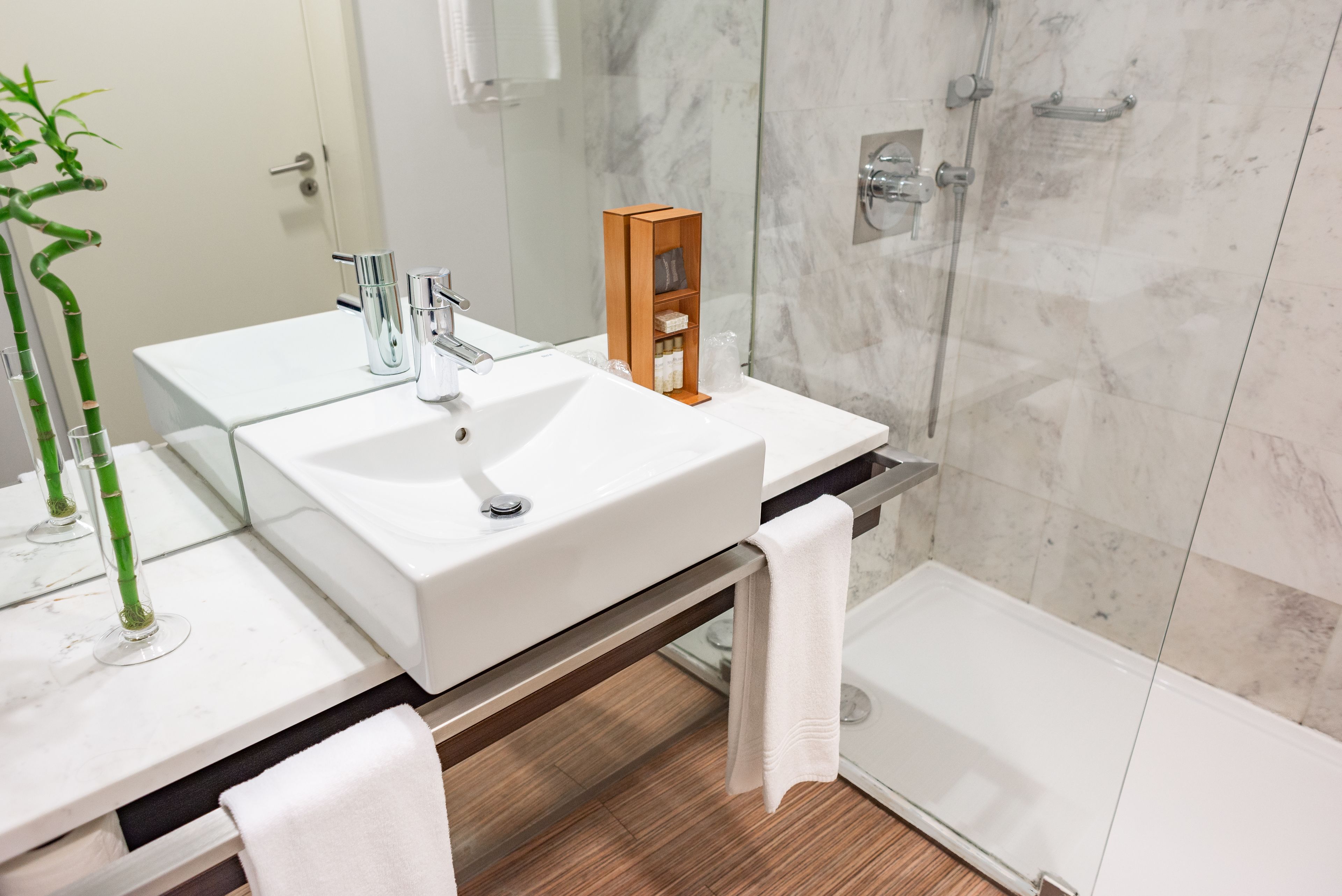Un bagno moderno con un lavandino bianco, uno scaffale in legno, uno spazio doccia in vetro e una pianta di bambù.