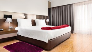 A modern hotel room with a large bed, a nightstand, a lamp, and a view of the city through large windows. - Hotel dos Zimbros (Sesimbra)