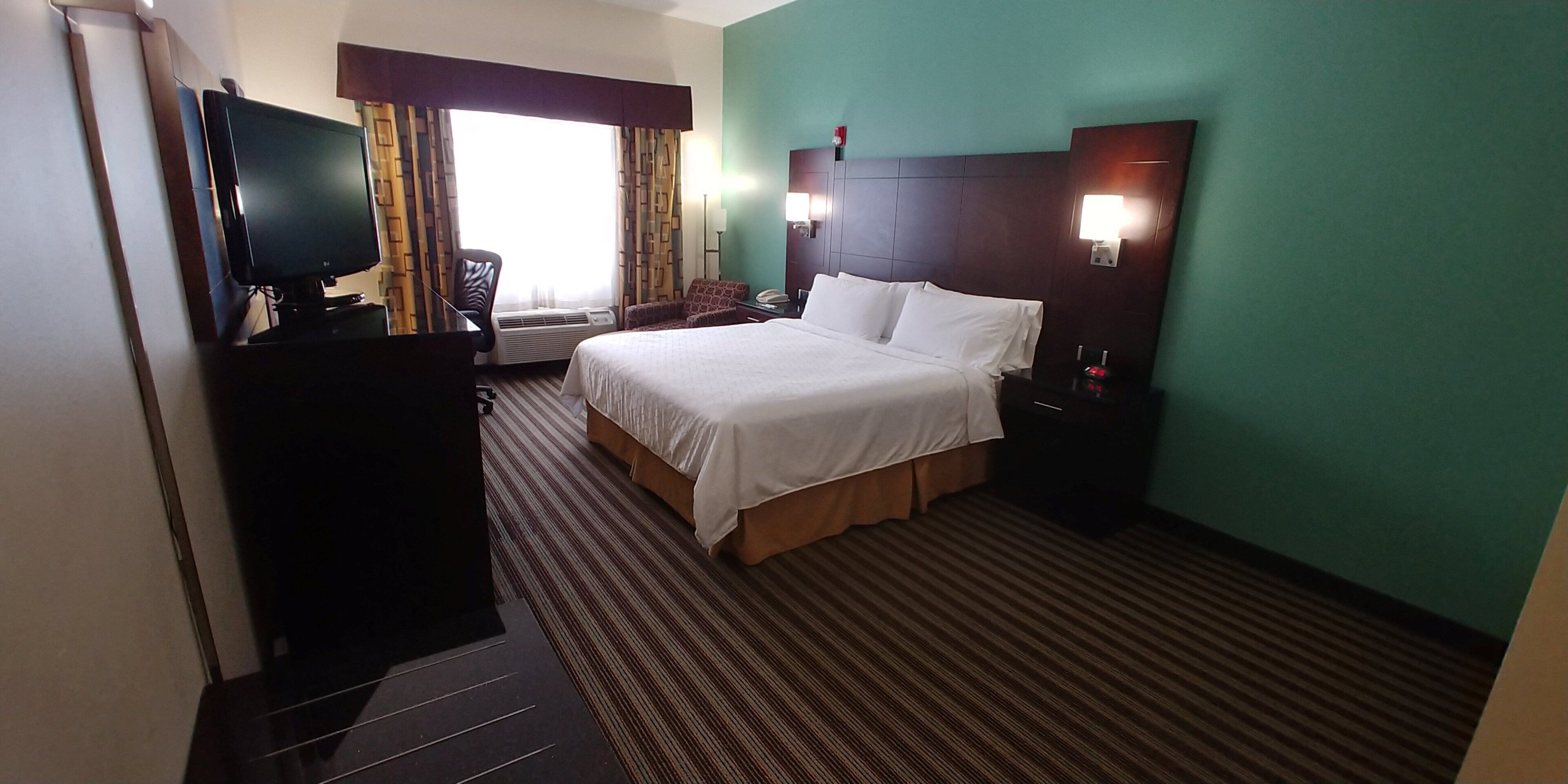 Habitación de hotel con cama, escritorio, silla, televisor y ventana con cortinas.