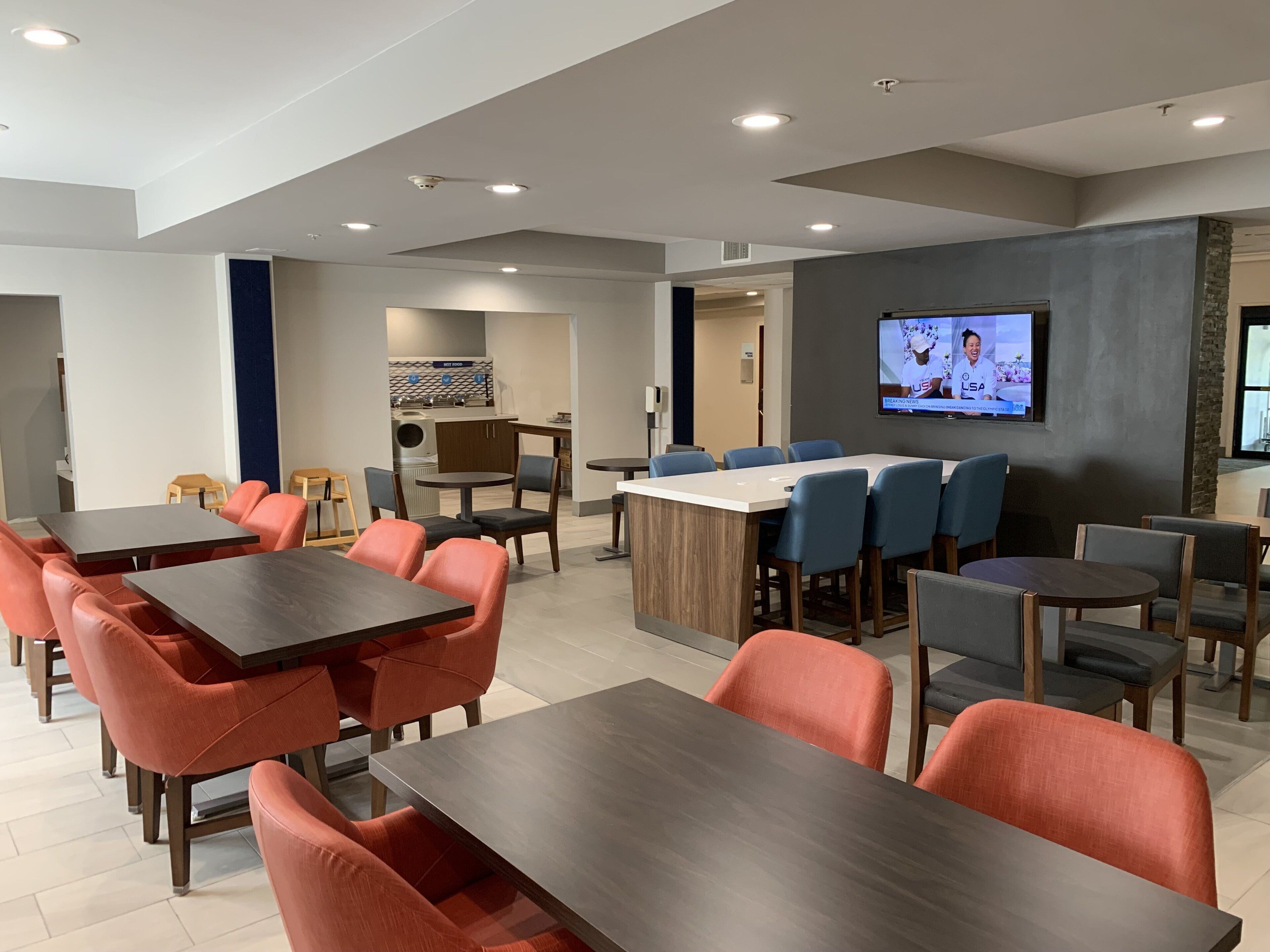 Un comedor de hotel moderno con varias mesas y sillas, una cocina al fondo y una pantalla de televisión mostrando un noticiero.