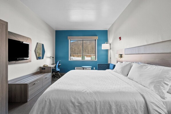 A hotel room with a large bed, a TV mounted on the wall, a desk with a chair, and a window with a view of trees.