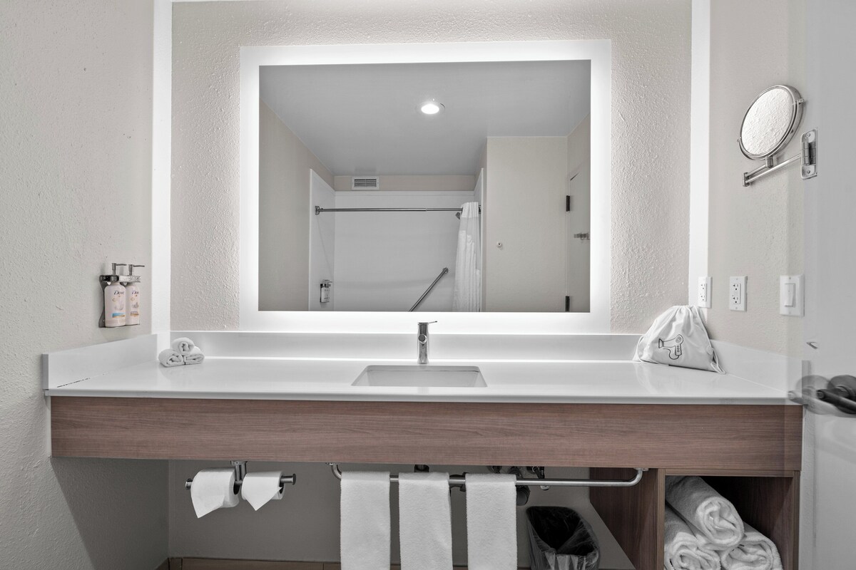 Een badkamer met een wit wastafelmeubel, een spiegel, een handdoekhouder en een douche zichtbaar in de spiegeling.