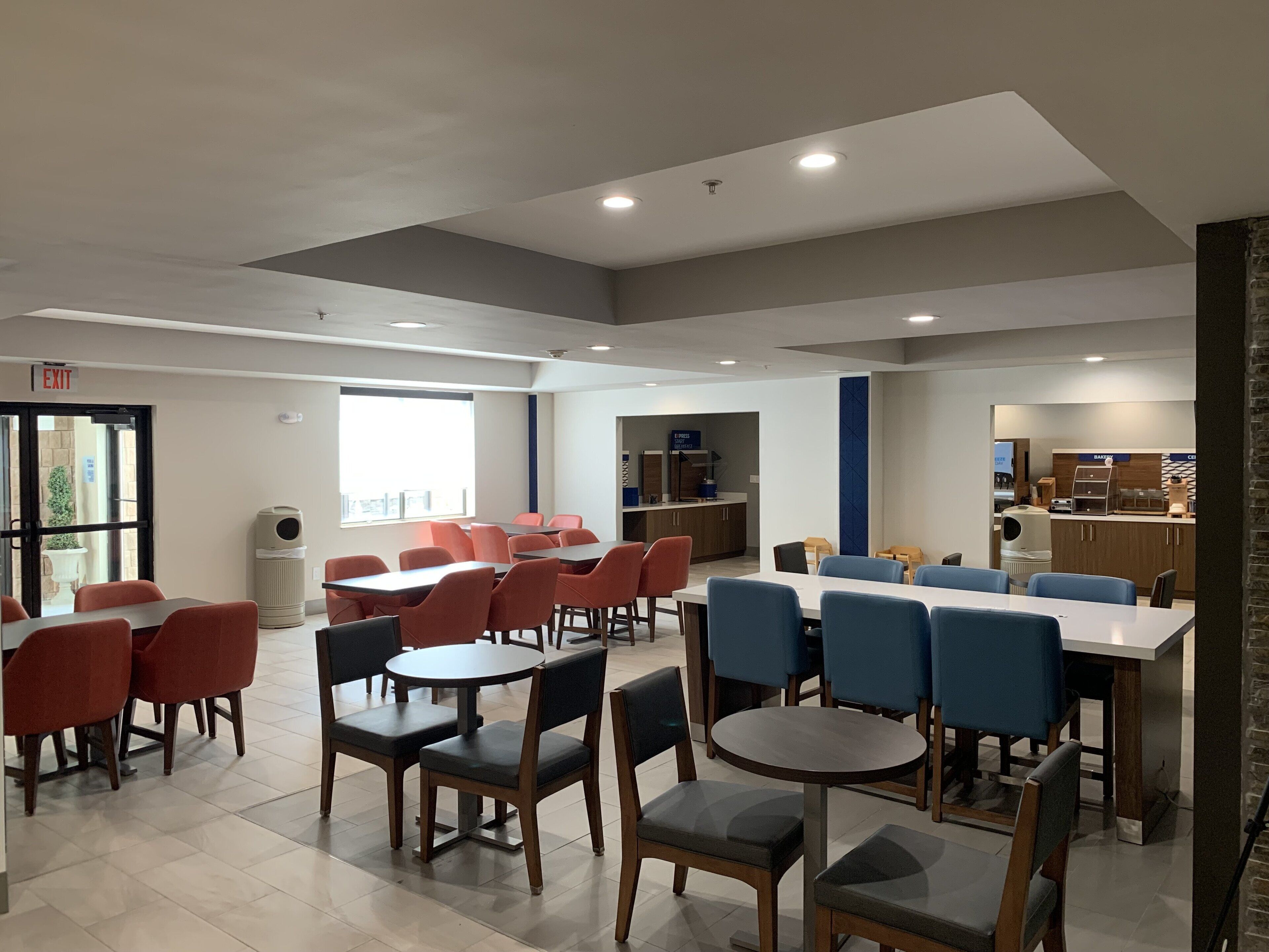 A modern dining area with tables and chairs, a kitchen area, and an exit sign.