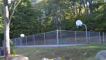 A fenced basketball court with a bench, surrounded by trees.