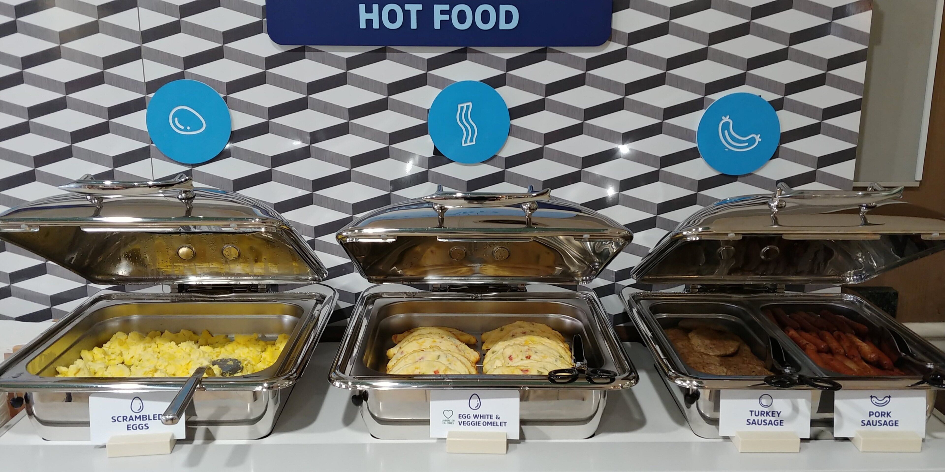 A buffet with labeled containers of scrambled eggs, egg white & veggie omelet, turkey sausage, and pork sausage.