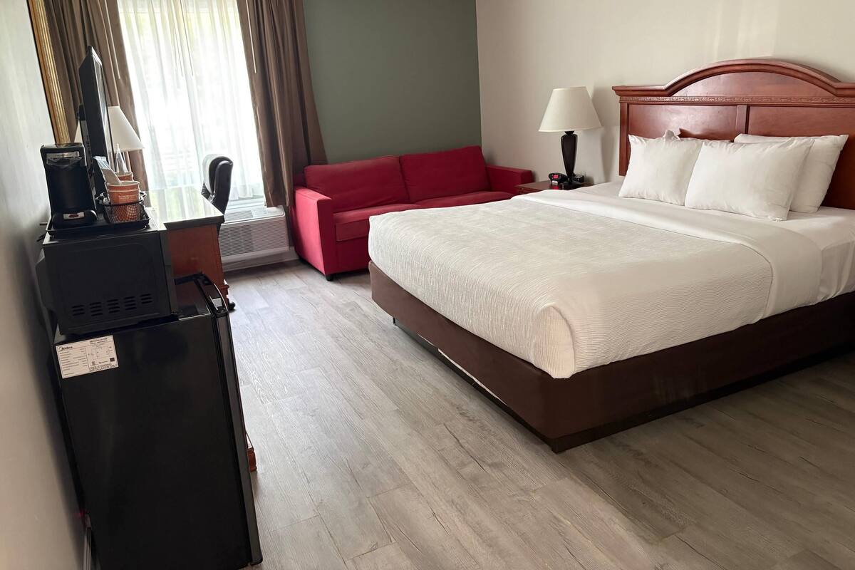 A hotel room with a bed, a red sofa, a lamp, a coffee machine, and a TV.