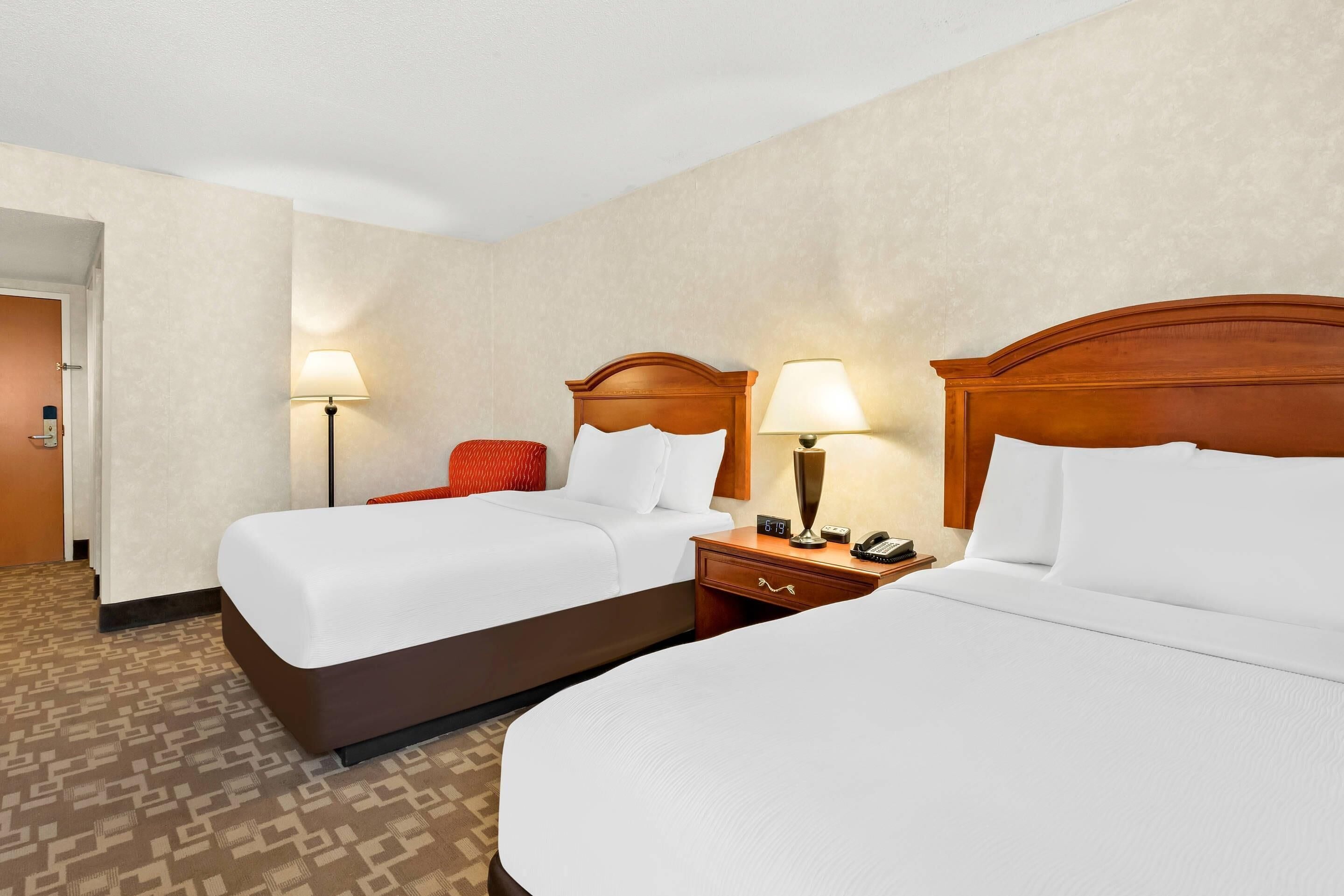 A hotel room with two beds, a wooden headboard, a nightstand with a lamp, and a door leading to another room.