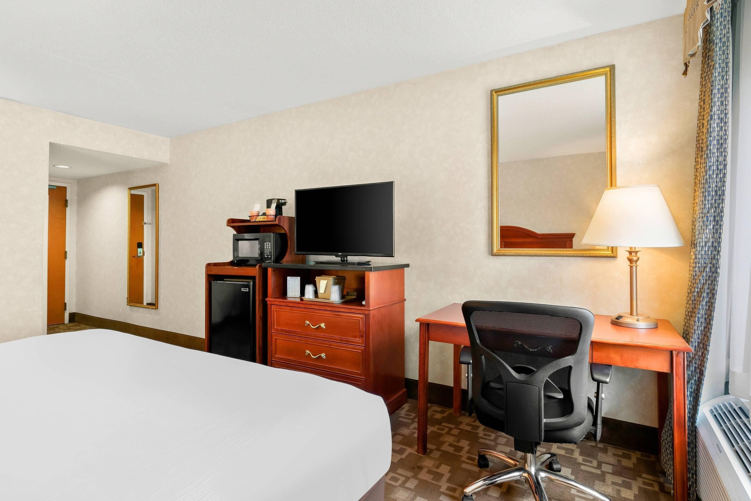 A hotel room with a bed, a desk with a chair, a television, a coffee maker, a mirror, and a lamp.