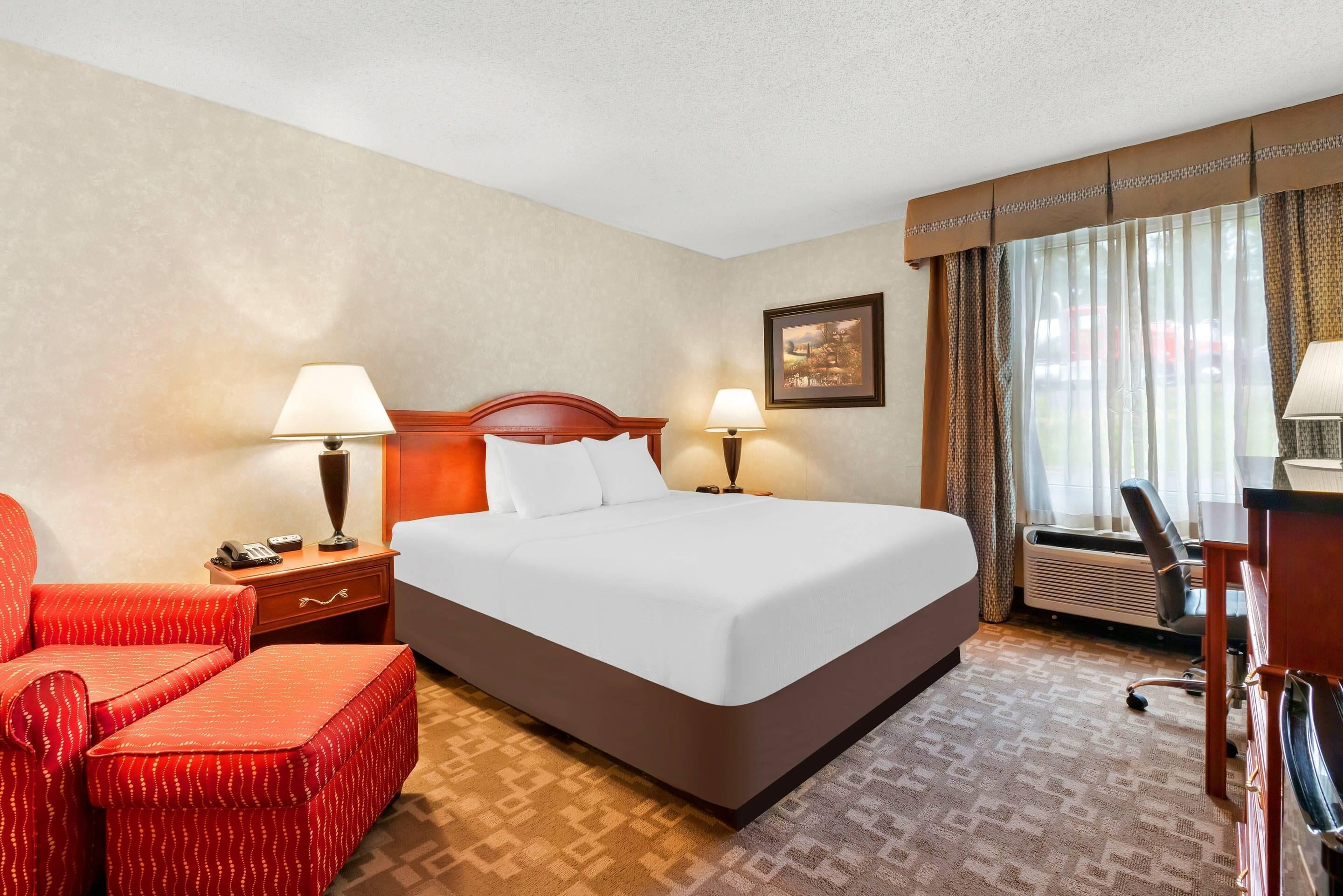 A hotel room with a large bed, two bedside lamps, a desk with a chair, a red upholstered chair, and a framed picture on the wall.