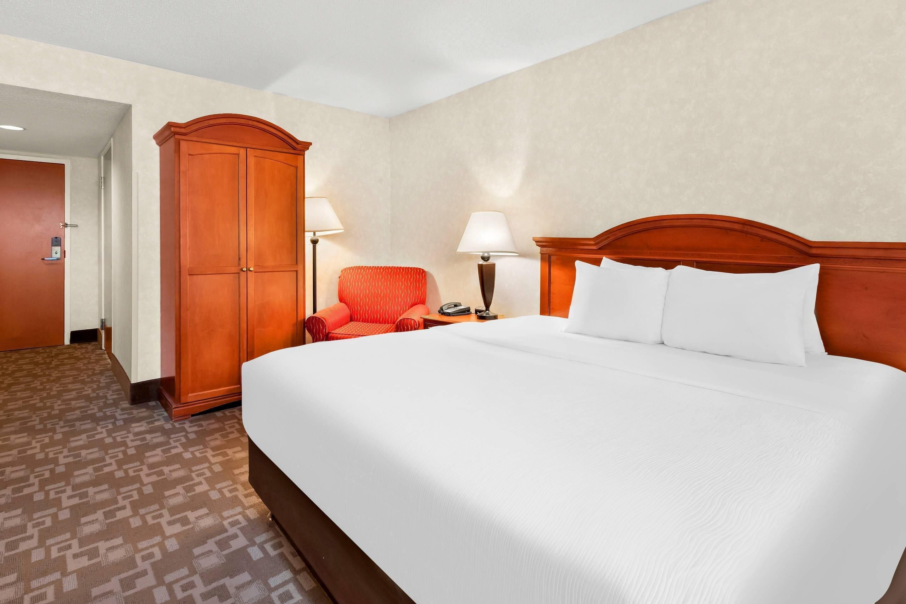 Habitación de hotel con una cama grande, un armario, un sillón y una mesita de noche con lámpara.