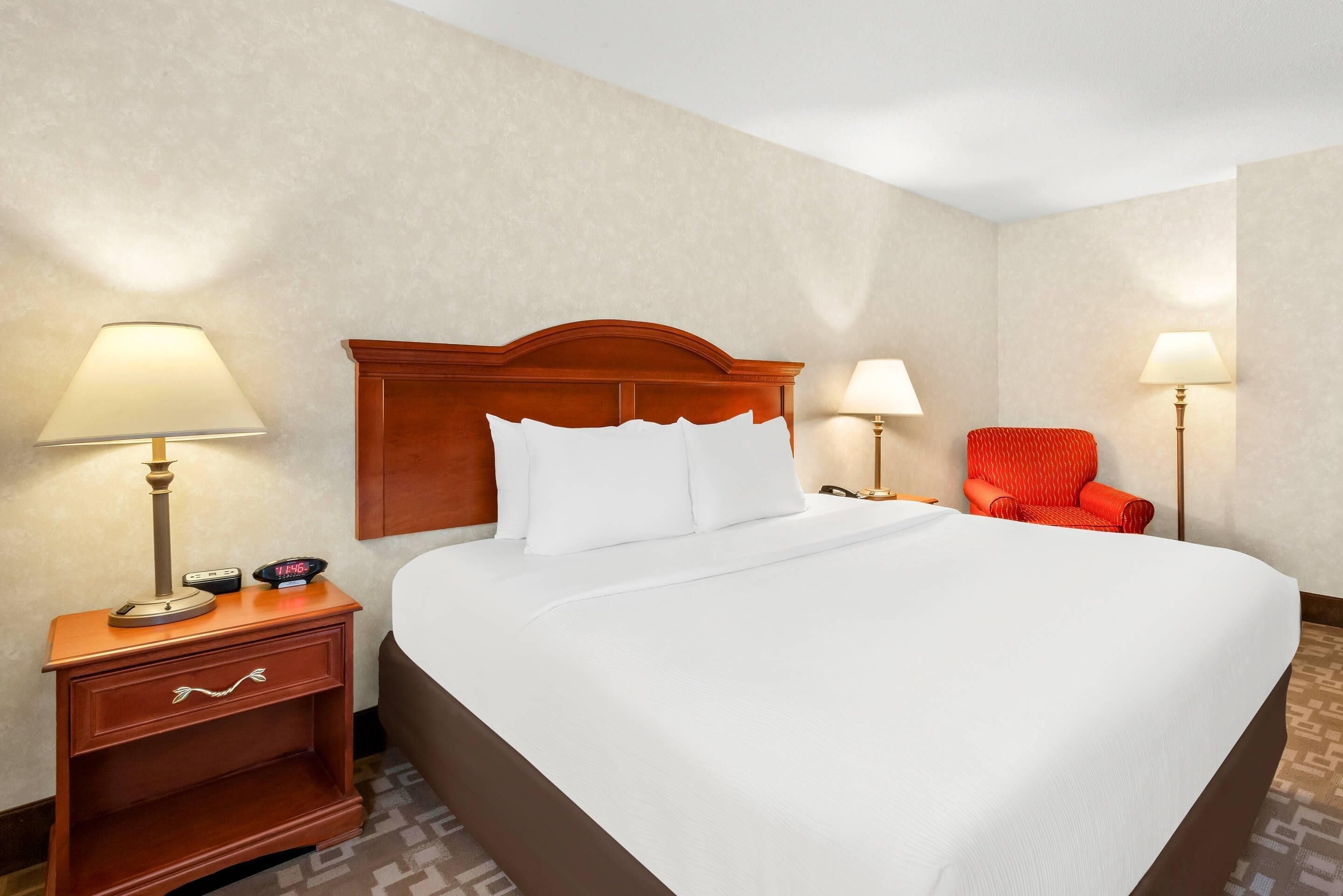 A hotel room with a large bed, two bedside lamps, a nightstand with a clock, and a red chair.