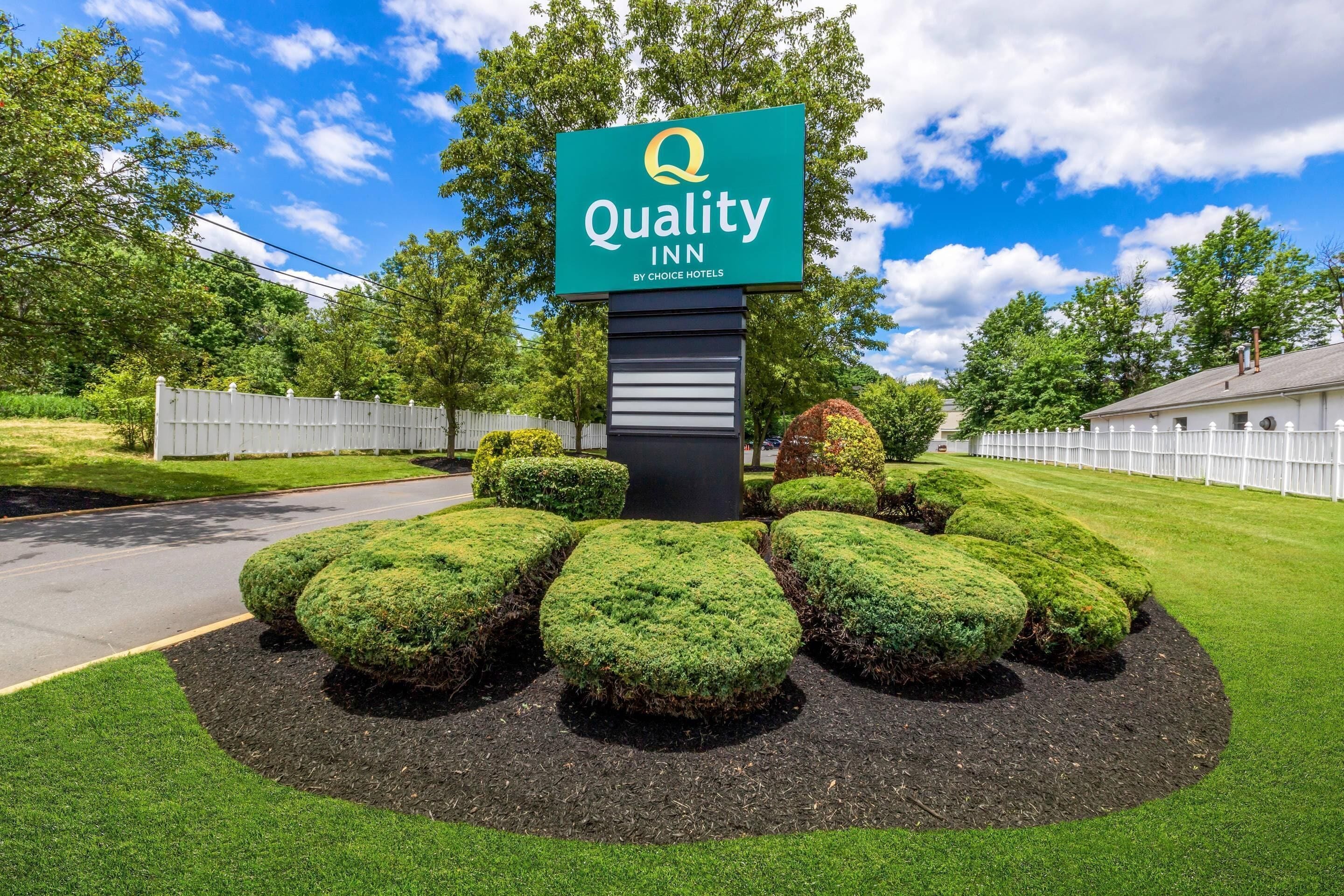 A Quality Inn sign with a logo, surrounded by well-manicured bushes and a clear sky.
