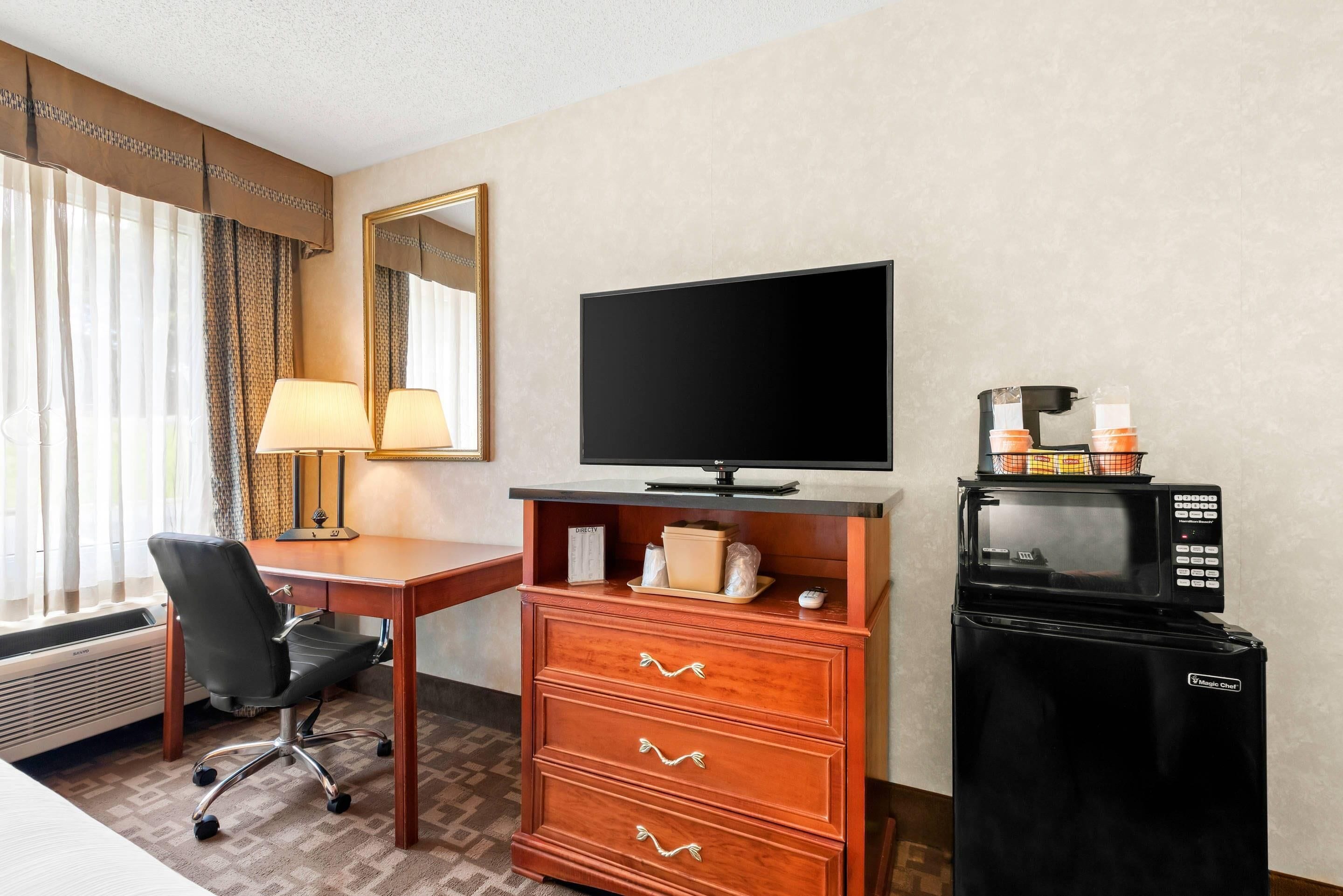A hotel room with a desk, chair, television, minibar, and a large mirror.