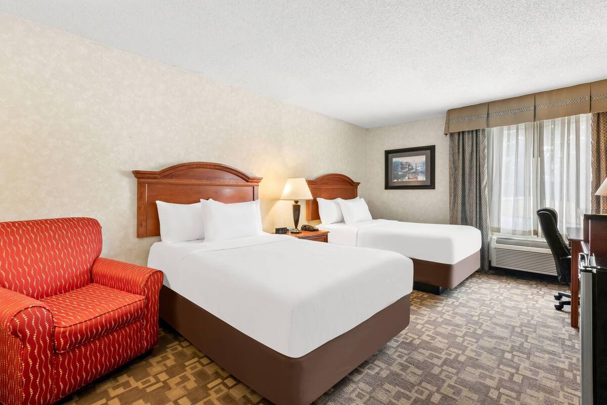 A hotel room with two beds, a red armchair, a desk, and a window with curtains.