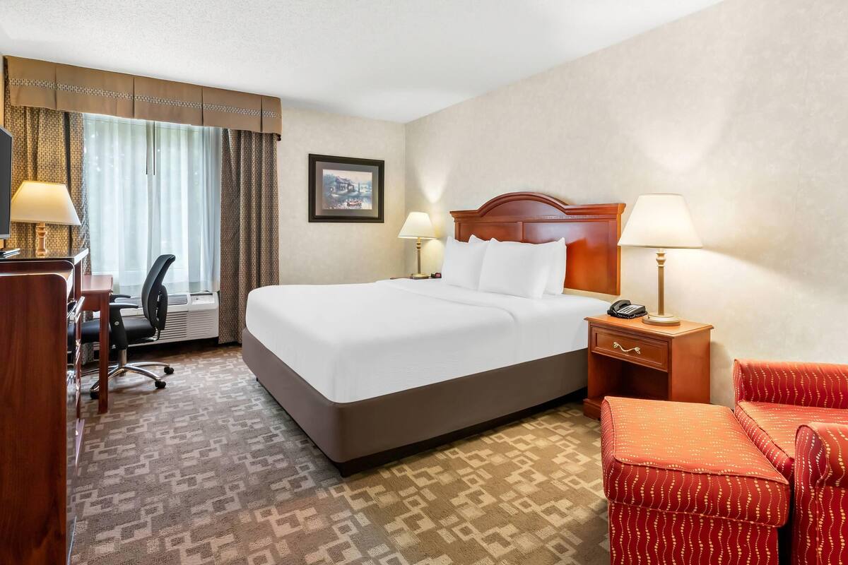 A hotel room with a large bed, two bedside tables with lamps, a desk with a chair, and a red upholstered chair.