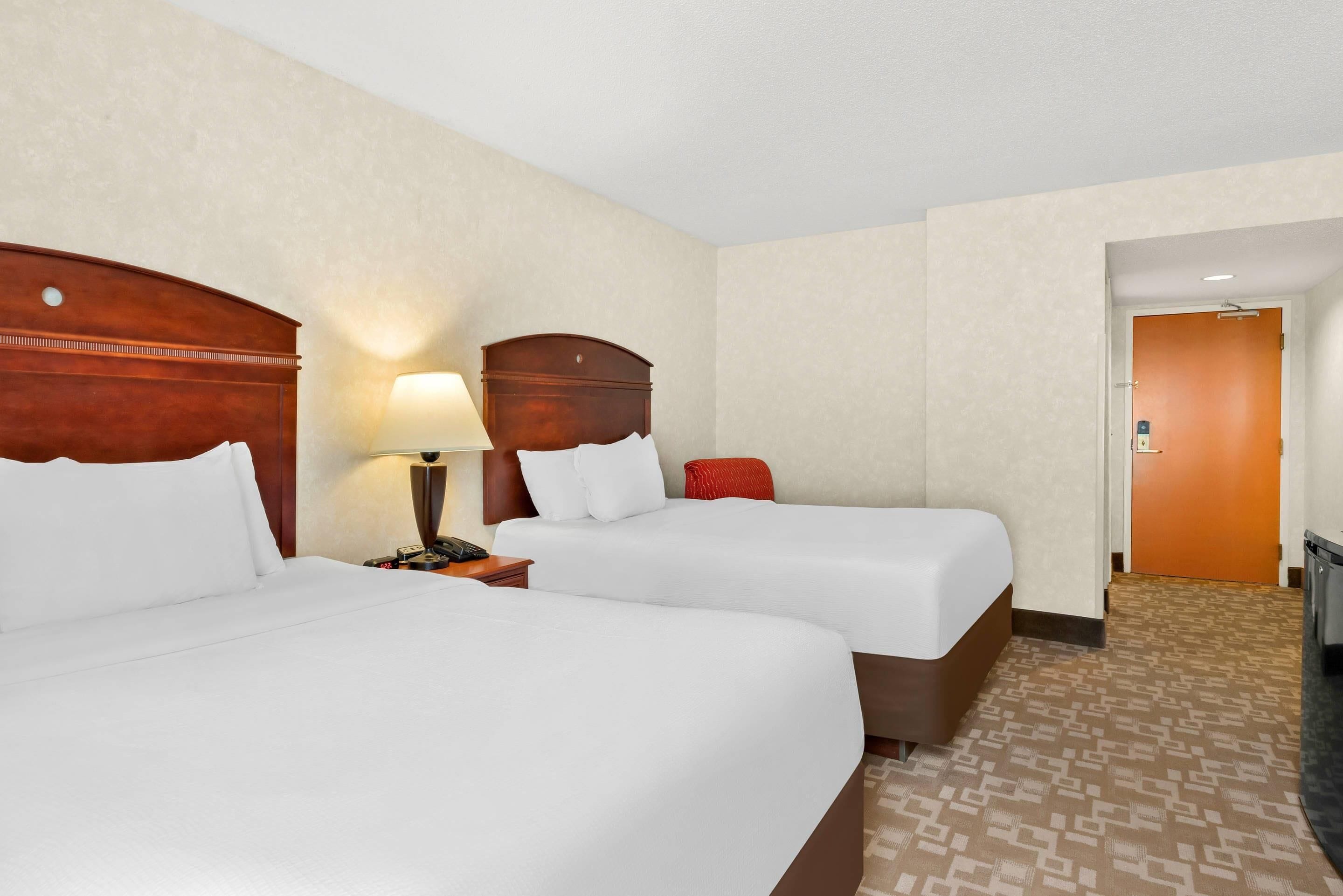 A hotel room with two beds, a nightstand with a lamp, and a door leading to another room.