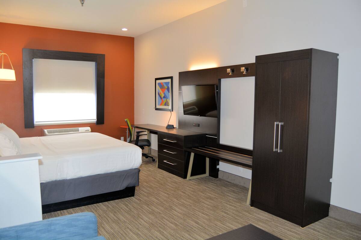 Ein Hotelzimmer mit Bett, Schreibtisch, Fernseher, Kleiderschrank und einem großen Fenster mit Jalousien.
