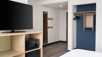 A hotel room with a flat-screen TV on a wooden shelf, a microwave, and a small refrigerator. There is a door with a mirror and a coat rack with hangers.