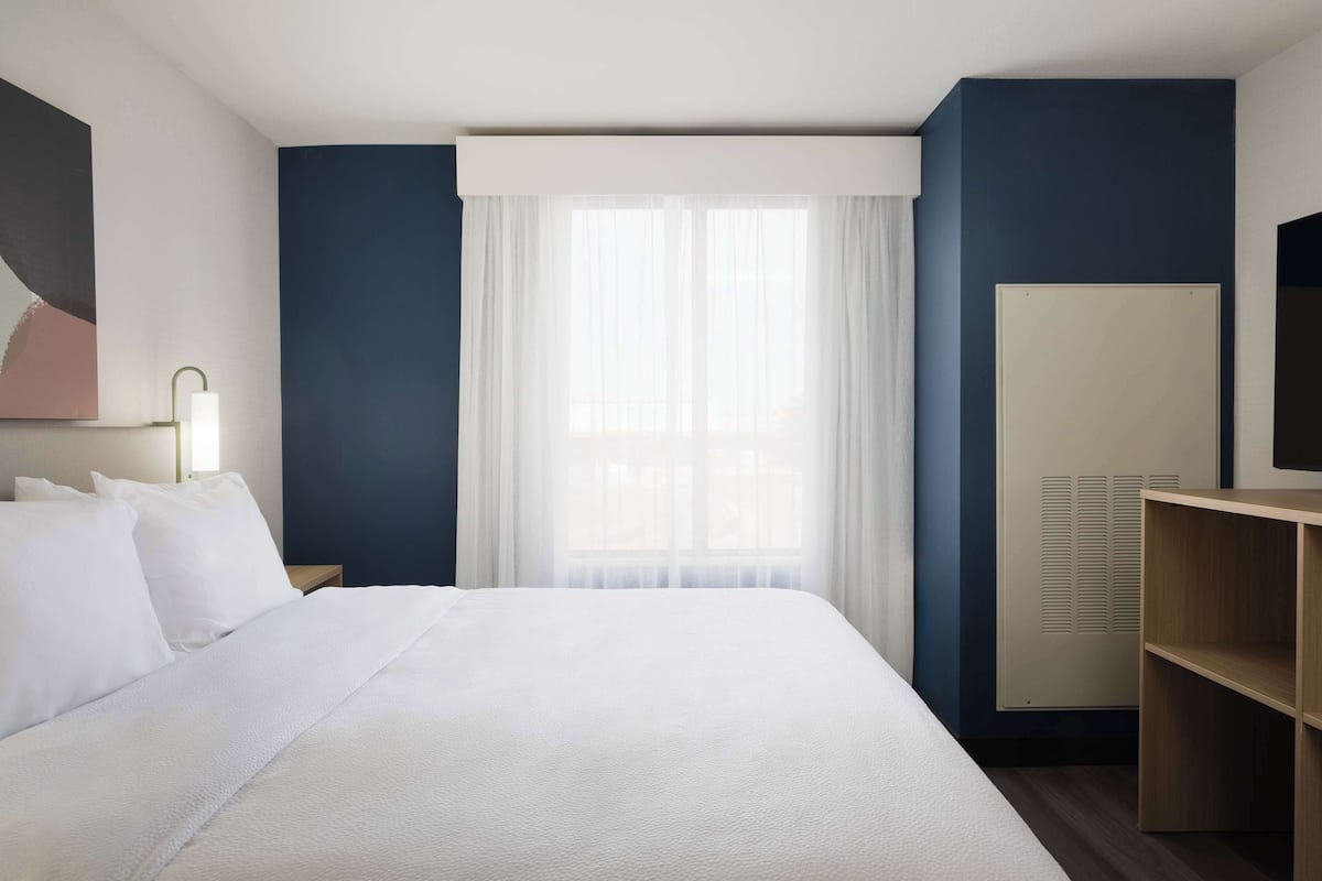 A hotel room with a large bed, a desk, a chair, a wardrobe, and a window with sheer curtains.