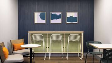 A room with a seating area featuring two cushioned chairs with orange cushions, a round table, and three white folding chairs against a wall with three framed artworks.
