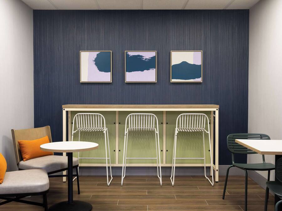 A room with a seating area featuring two cushioned chairs with orange cushions, a round table, and three white folding chairs against a wall with three framed artworks.