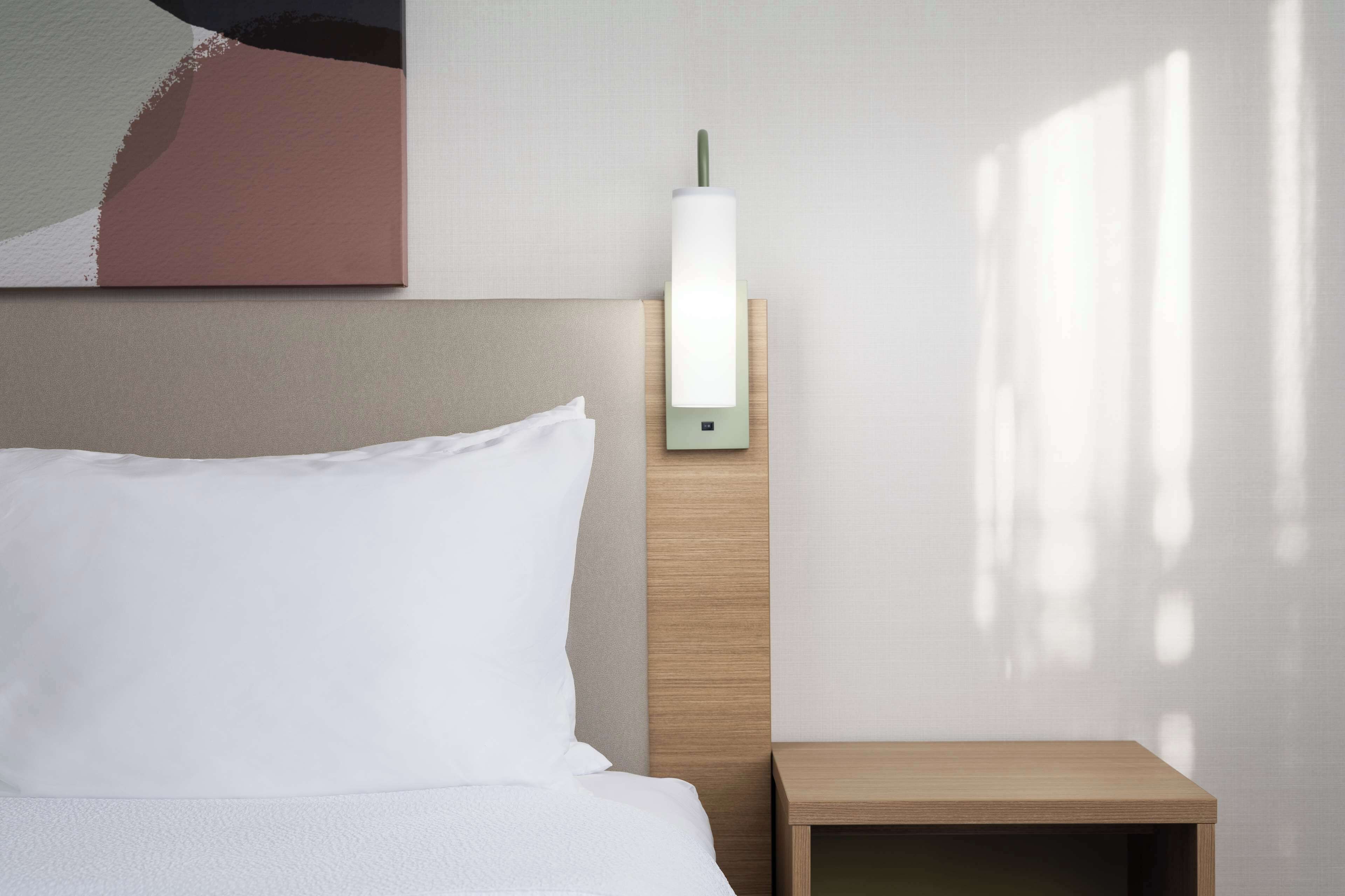 Un lit bien fait, avec du linge de lit blanc, une table de chevet et un luminaire fixé au mur.