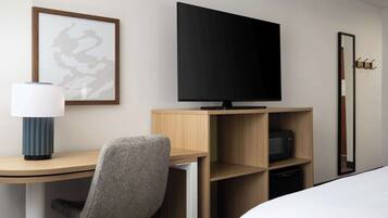 Quarto de hotel com cama, escrivaninha, cadeira, televisão, micro-ondas e quadro na parede.