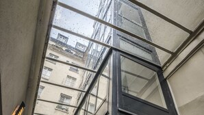 A view through a glass door with a metal railing, leading to a building with multiple windows and a visible fire exit sign. - Chabrol Opéra (Paris)