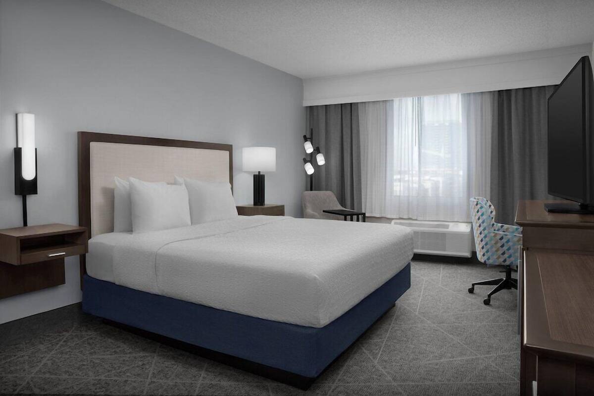 A hotel room with a large bed, bedside tables, a desk, a chair, a TV, and a window with curtains.