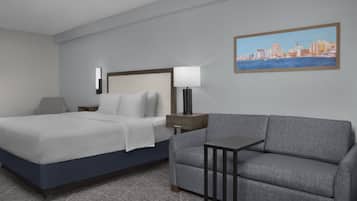 Habitación de hotel con una cama, un sofá gris, una mesita de noche, una lámpara y una pintura de un paisaje urbano en la pared.