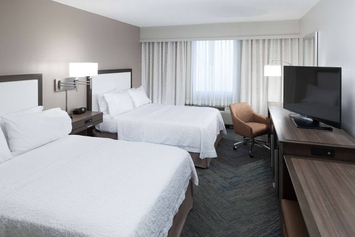 A hotel room with two beds, a desk with a TV, a chair, and a window with curtains.