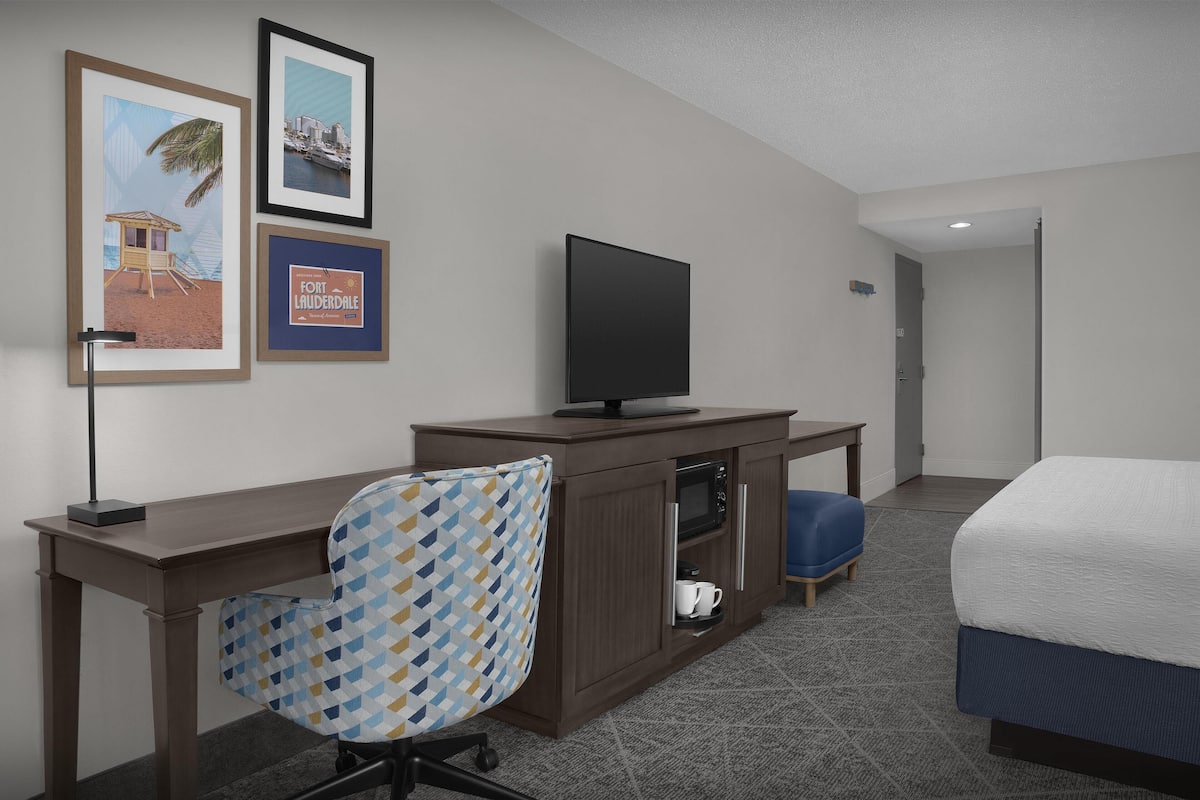A hotel room with a bed, a desk with a television, a chair, a cabinet, and framed pictures on the wall.