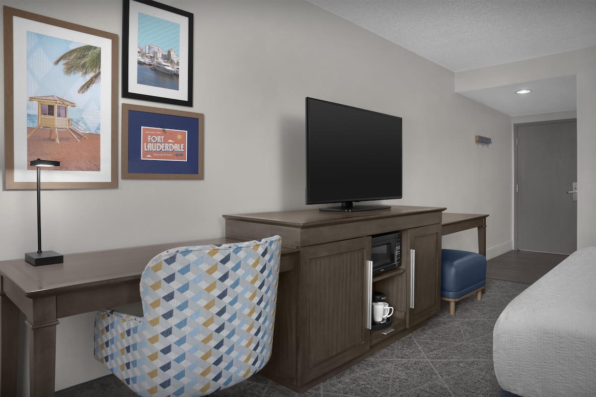 A hotel room with a wooden desk, a television, a patterned armchair, a minibar, and framed pictures on the wall.