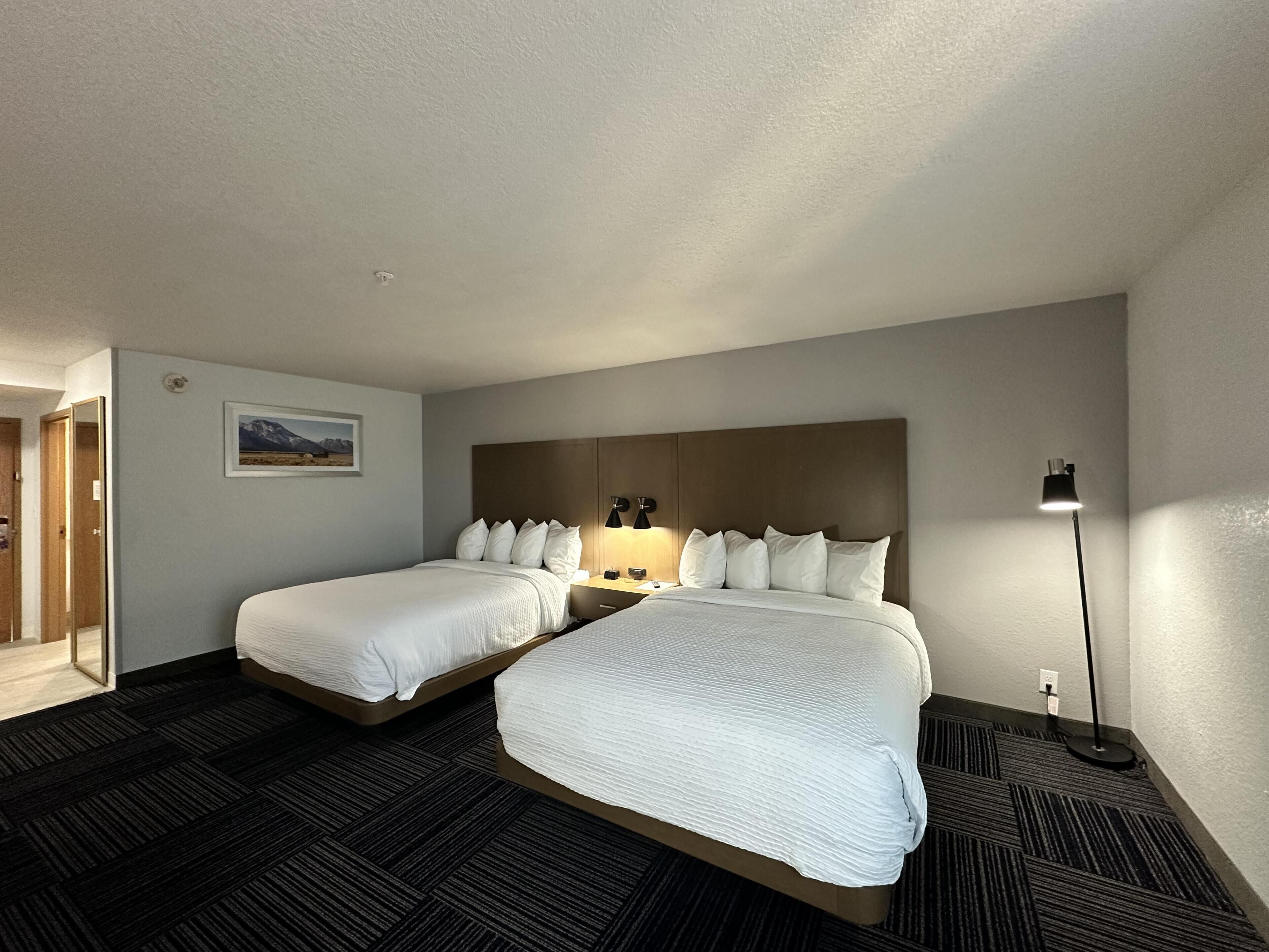 Habitación de hotel con dos camas, una mesita de noche, una lámpara, un cuadro en la pared y suelo cubierto de alfombra.