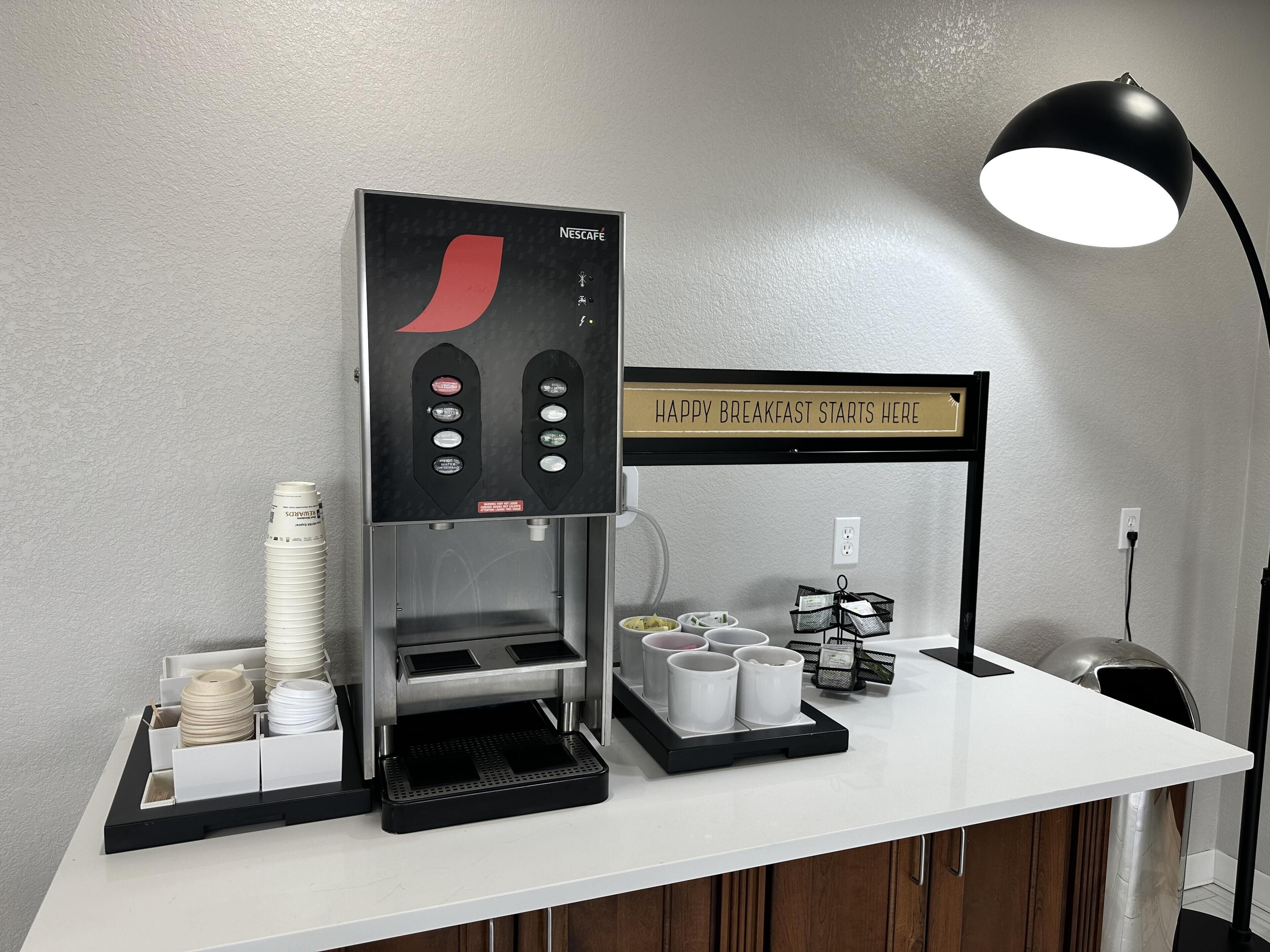 Uma máquina de café com um letreiro que diz 'HAPPY BREAKFAST STARTS HERE' e uma luminária em uma mesa.