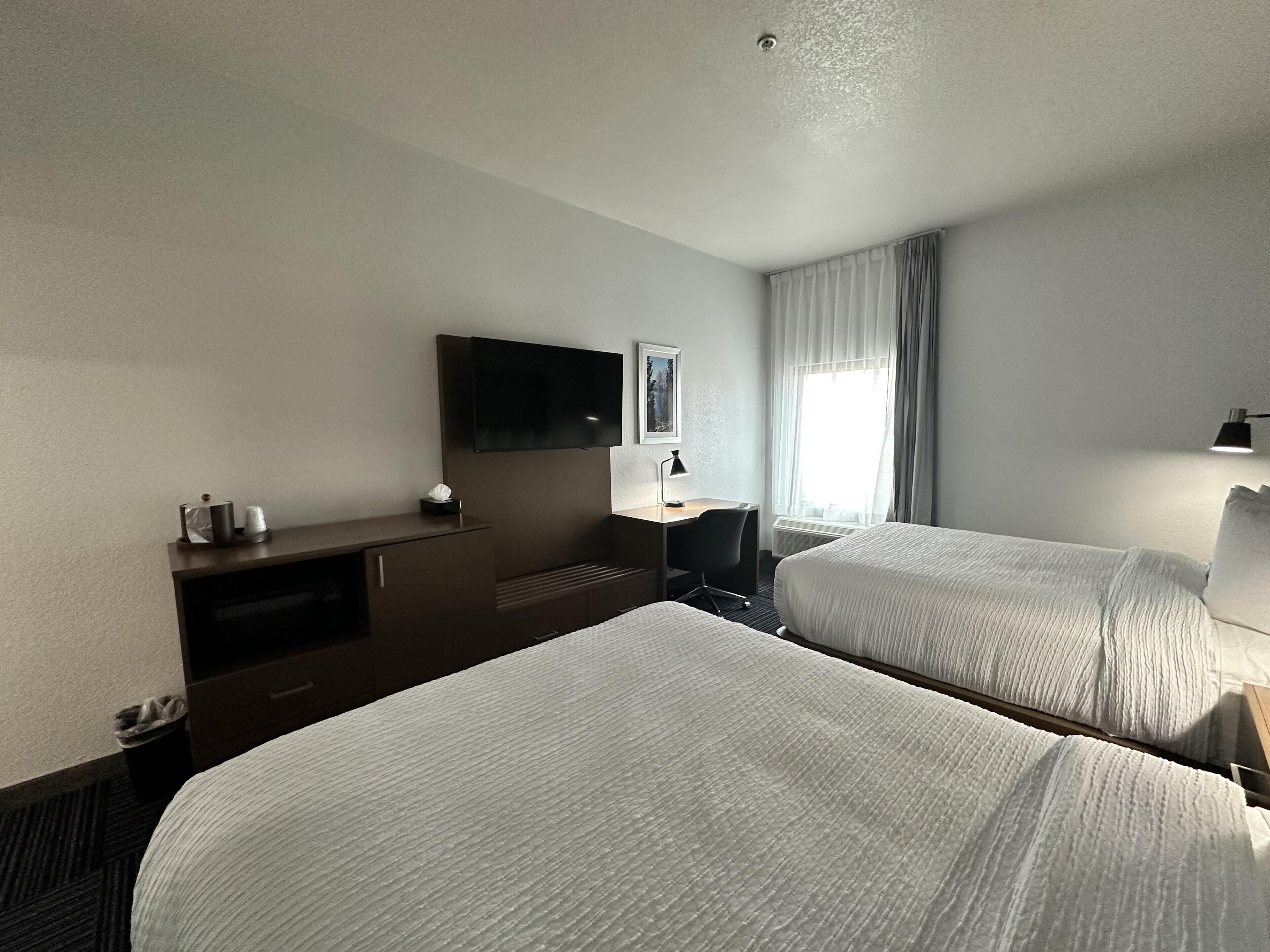 A hotel room with two beds, a desk, a TV, and a window with curtains.